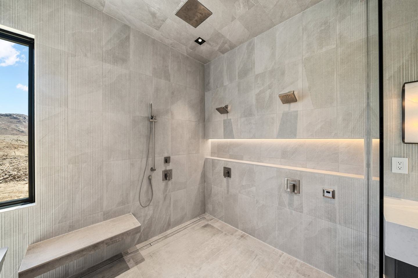 Walk-in stone shower with dual heads, lit niche, and a desert view window