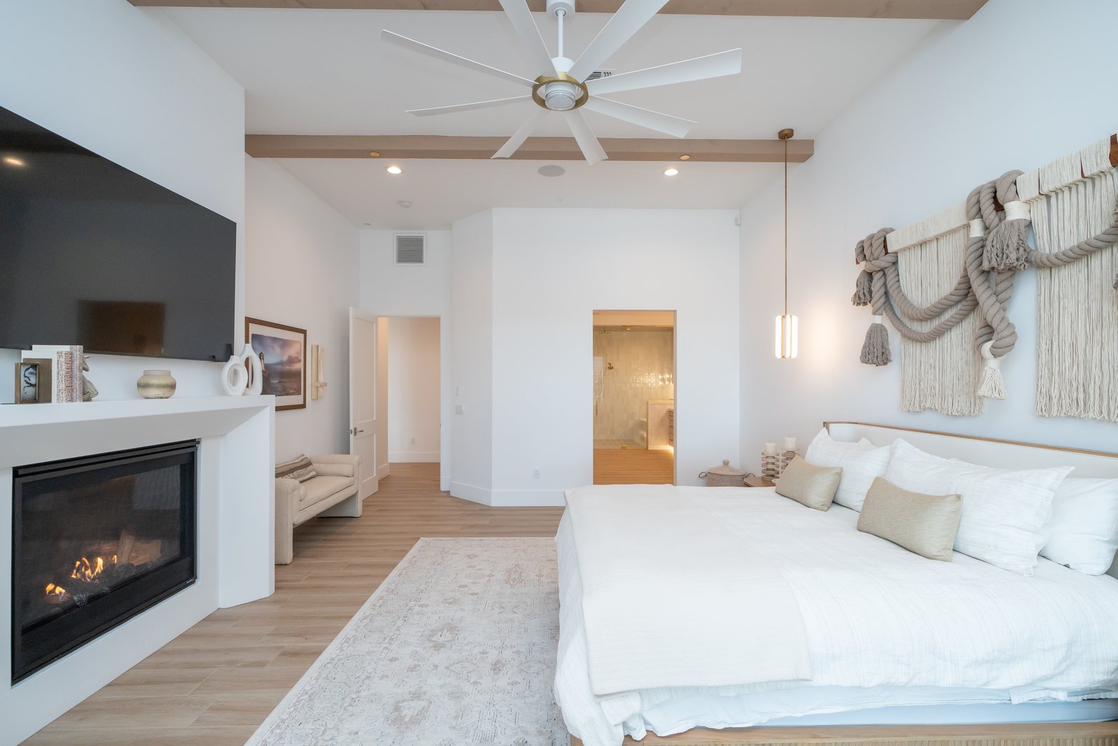 Primary suite with a linear fireplace, beamed vaulted ceiling, woven fiber art wall, and ensuite beyond