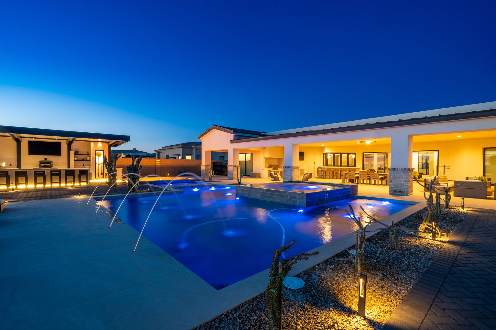 Backyard pool and spa at dusk with covered patio, outdoor kitchen, and desert landscape lighting