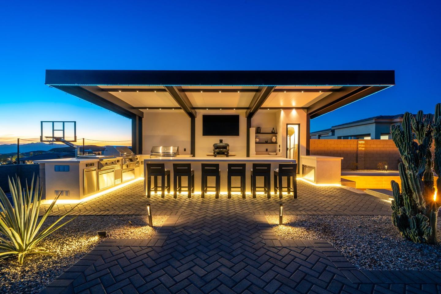 Covered outdoor kitchen and bar at dusk under a cantilevered ramada with integrated lighting
