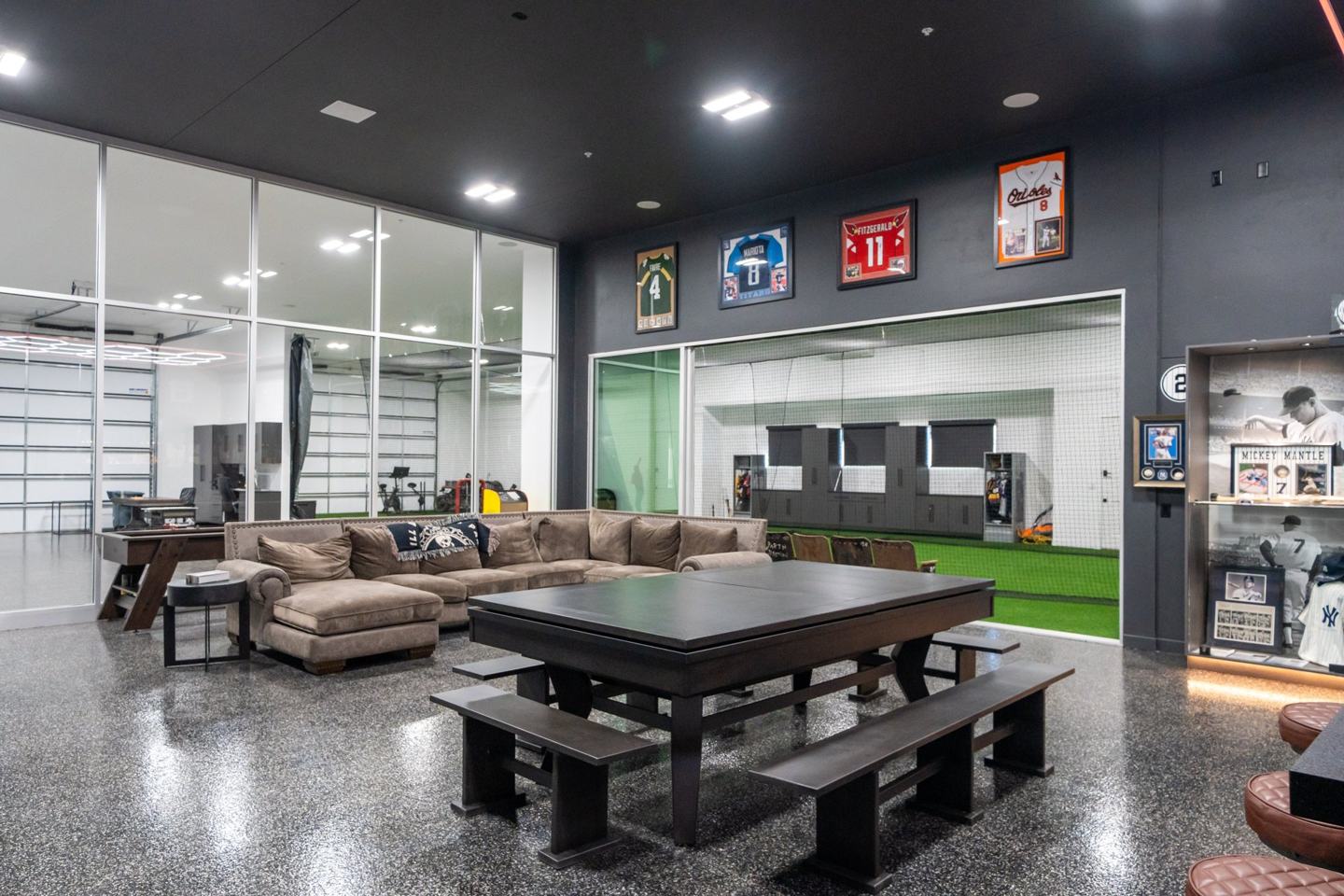Sports lounge with framed jerseys, sectional seating, and a window into the batting cage